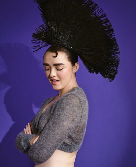 Maisie Williams standing with her left shoulder facing the camera, wearing a cropped grey knitted top and an extravagant black headdress with her arms folded across her body, against a deep-purple background