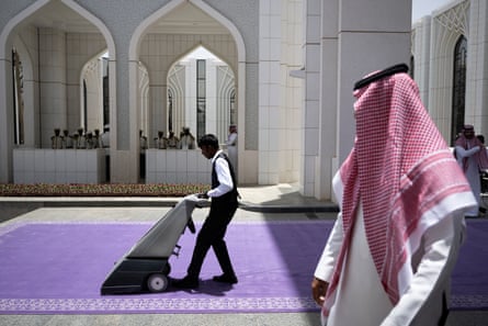 A man in a uniform vacuums a purple carpet in the courtyard of a white building. A man wearing a keffiyeh and white robe is in the foreground