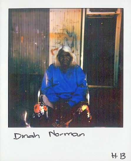 Dinah Norman est assise sur une chaise devant une vieille maison.  Elle porte un pull bleu et un pantalon à motifs