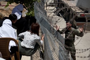 Israeli soldier in fatigues holds his hands out wide as a group of people talk to him through a metal fence topped with barbed wire