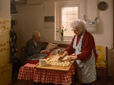 A woman stands at a kitchen table cutting dough into shapes while her husband sits reading a newspaper
