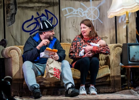 Robertson and Dow Blyton, holding a baby, sit on a sagging sofa in front of a graffitied wall, with a standard lamp and TV to one side.