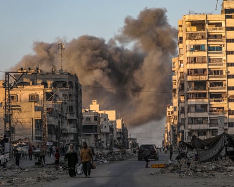 Smoke billows following an Israeli airstrike on Gaza City, while a man and woman walk away carrying a plastic bag