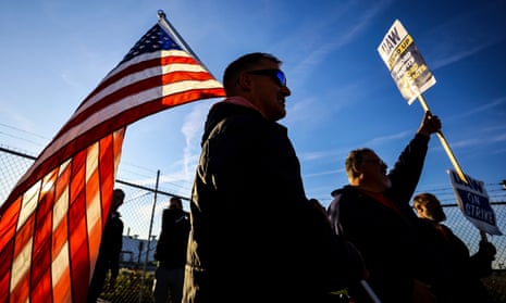White men holding picket signs are backlit as the sun picks up the red-white-and-blue flag held by one, beneath a blue sky.