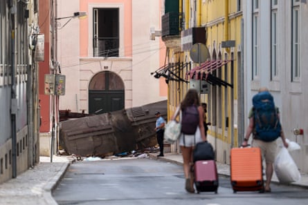 Tourists wearing backpacks and pulling suitcases walk down a street, at the foot of which the wreckage of the streetcar is visible