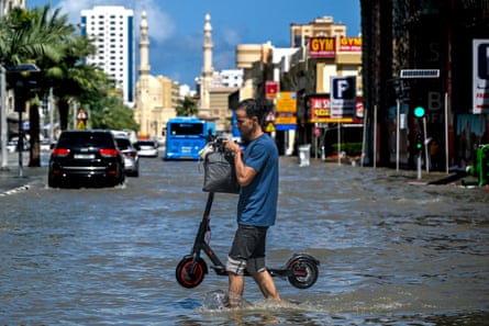 A young man with T-shirt and shorts holds a scooter above water as he wades across a street ankle-deep.