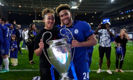 Lauren, in street clothes, and Reece, in his blue Chelsea uniform, pose for a picture on the pitch, each holding one handle of a very large silver trophy