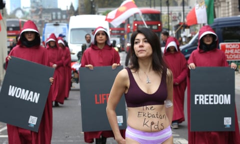 A woman in her underwear walks in front of women dressed in costumes from The Handmaid's Tale carrying sighns that read 'Woman, Life, Freedom'