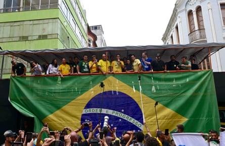 A man flanked by people in Brazil’s football colours addresses a crowd of photographers while standing on a raised platform above a gigantic Brazilian flag.