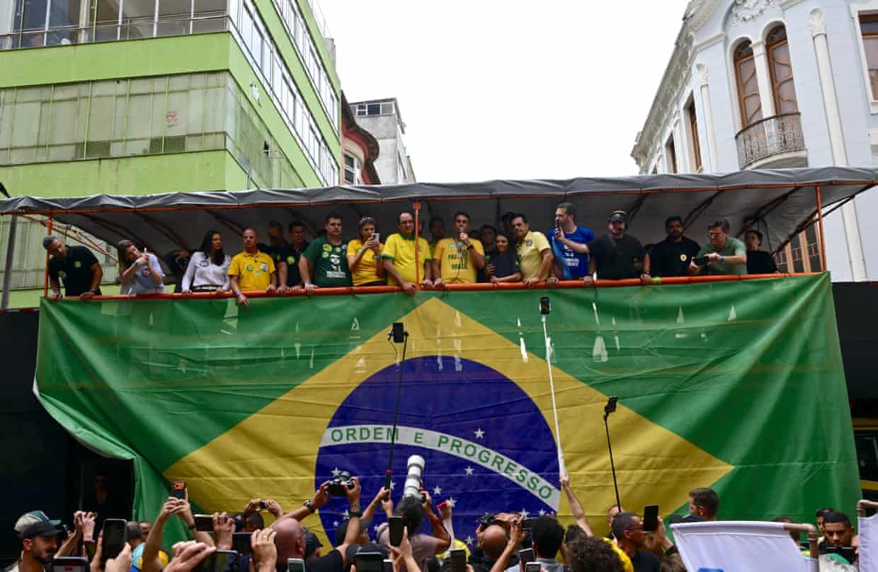 A man flanked by people in Brazil’s football colours addresses a crowd of photographers while standing on a raised platform above a gigantic Brazilian flag.
