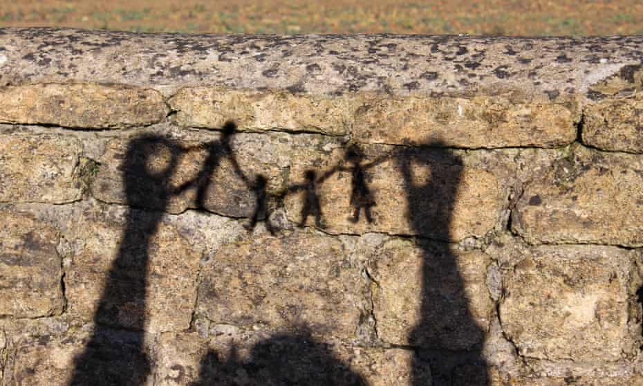 Photo showing a family paper chain of people (father, son, daughter and mother), cut out of white paper. The people paper chain is pictured being held with two hands / fingers in the strong afternoon sunshine, with the shadows of the people appearing as clear black silhouettes against a cobblestone wall.