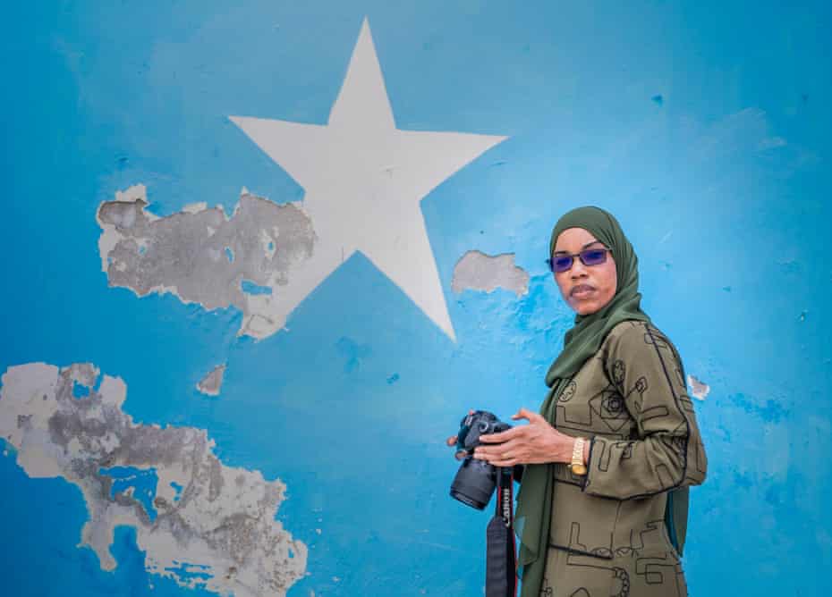 A woman in headscarf and carrying a camera in front of a blue wall with a big star