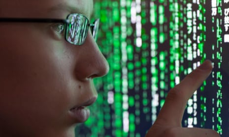 A young man with glasses is watching futuristic symbols on a computer screen. Symbols are reflecting in the man’s glasses.