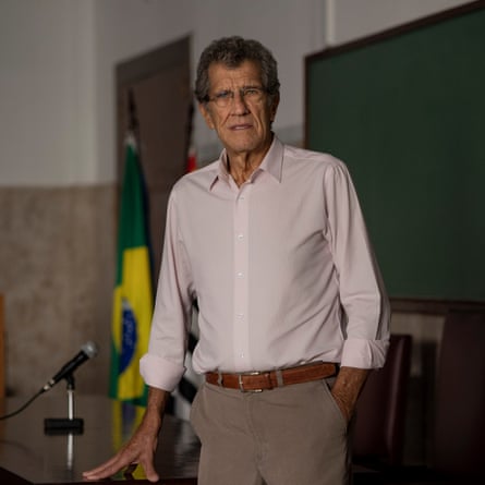 An older white man with a lined face and a serious demeanour stands at a desk in a lecture hall with the Brazilian flag behind him