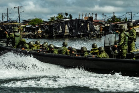 A boat carrying members of the navy, in uniform and holding guns, passes a row of rundown houses on the shore