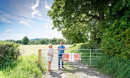 A couple stand next to a banner attached to a fence that reads:”No Pylons” outside a field where a pylon is planned to be built