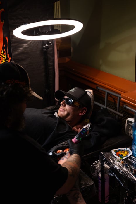 A man in sunglasses and baseball cap lies under a circular fluorescent lamp while a tattooist works on his arm