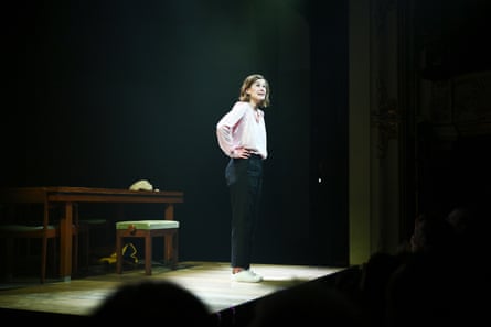 she is standing on a darkened stage with the spotlight on her. She wears a pale silk blouse and dark trousers.