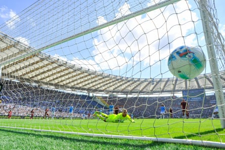 A camera behind the goal captures Lorenzo Pellegrini’s shot for Roma hitting the net against Lazio underneath a blue and sunny sky.