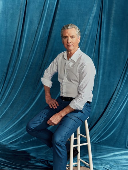 Gavin Newsom sitting on a tall white stool at a slight angle to the camera, looking straight at it, his right hand resting on his right thigh and his left wrist resting on his left thigh, wearing a white shirt with the sleeves rolled up and no tie, against a turquoise velvet background
