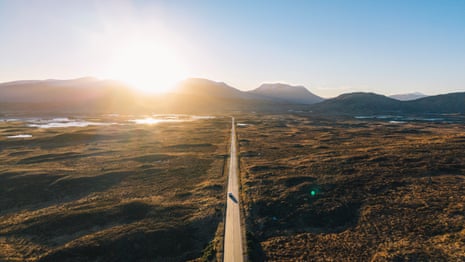 A straight road cuts through moorland towards distant mountains, with a single vehicle visible