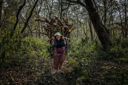 A woman with a large bundle of wood on her back smiles in a forest setting