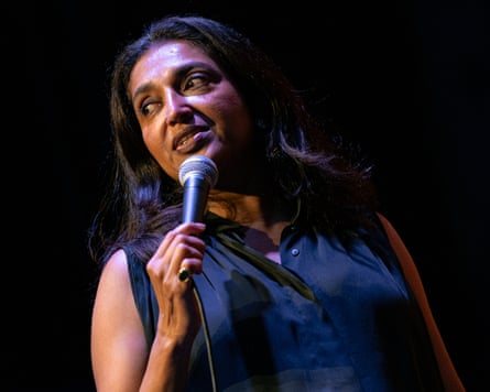 Comedian Sindhu Vee holds a microphone, looking off to the side under stage lighting
