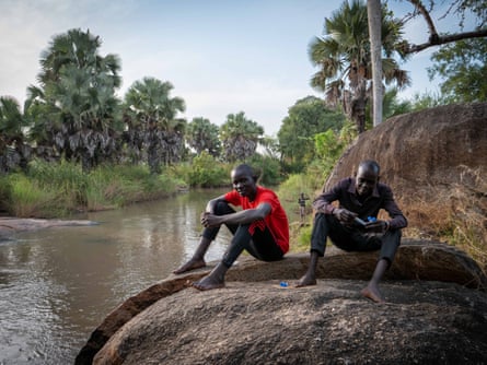 A young man and an older man, who is holding some test tubes, sitting on a big rock by a tropical river with palm trees along it