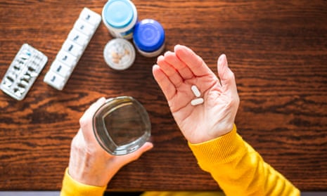 Hands of an older woman in a nubbly jumper holding a glass of water and two pills against a table with two jars of pills, a round pillbox and a dosette box with days of the week printed on its lids