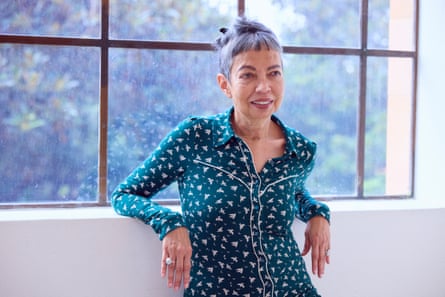 Michelle de Kretser wearing a green pattered top, standing in front of a window with trees in the background outside.
