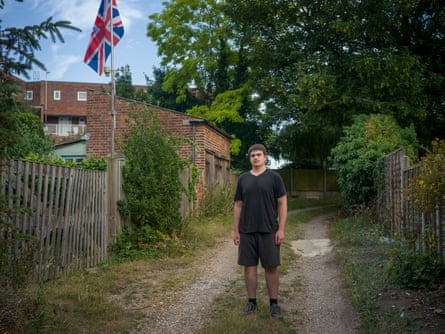 A young man stands in a lane next to a house with a union jack flag flying