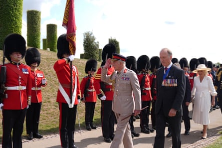 King Charles in a tan military suit salutes a royal guard in the grounds of the National Memorial Arboretum