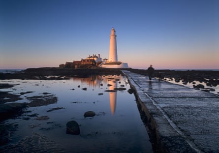 Causeway leading to a lighthouse at dusk in winter with reflection in the water. A man is walking on the causeway