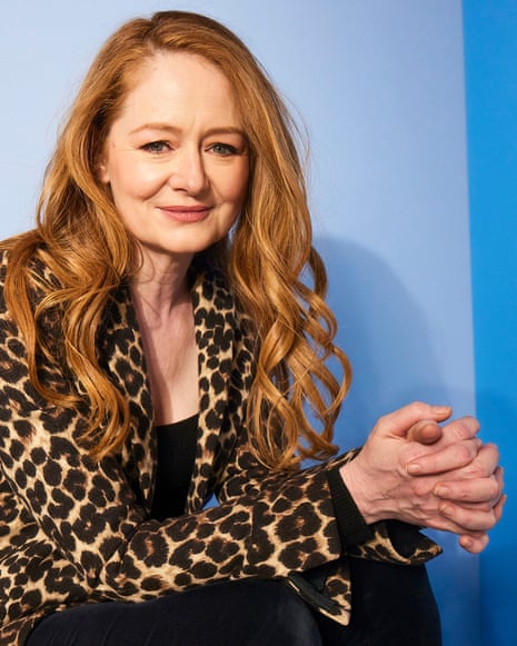 Miranda Otto with long auburn hair and wearing leopard print blouse, photographed against a blue wall, smiling.