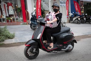 A woman driving a moped wearing a black facemask with a dog in a bag sitting on her lap.