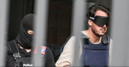 A young man, in armoured vest and blindfold, accompanied by a police officer in a balaclava, photographed while in custody in 2022.