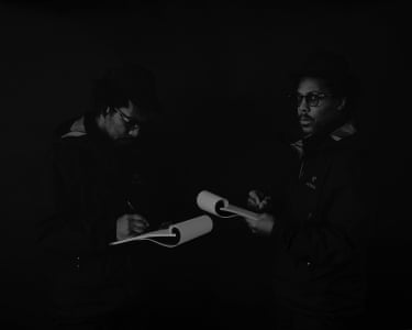 dimly lit black and white photo of two men facing each other holding notepads