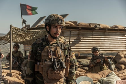 A soldier in army fatigues with a big walkie talkie and other soldiers and a flag behind him