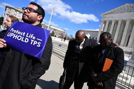 A person holds a sign that reads ‘protect life uphold TPS’ as two men behind him bow their heads in prayer.