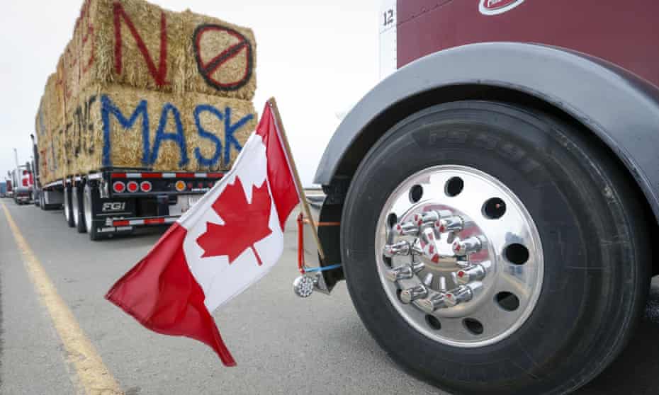 The Canadian flag is reflected in a wheel hub of a tractor-trailer that is part of a truck convoy protesting Covid-19 safety restrictions. Ahead of this truck, there is another tractor trailer hauling bales of hay or wood that are spray painted with the slogan "NO MASK" in capital letters.