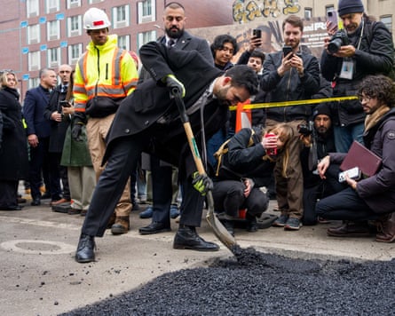 Zohran Mamdani, wearing a suit and coat, filling a pothole