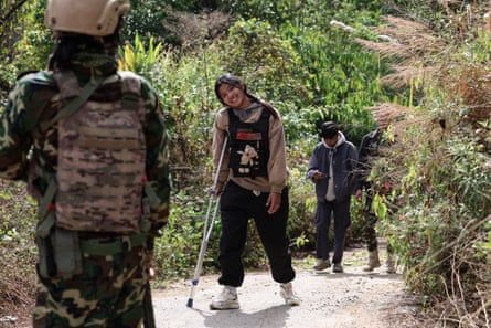 A girl in tracksuit bottoms and bulletproof vest walks along a rural lane using a crutch