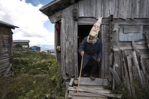 An elderly man with a walking stick emerges from a wooden hut wearing a tall furry headdress with a face painted on it