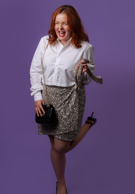 Lucy Edwards standing on her right leg, with her left leg bent at the knee, laughing at the camera, wearing a white, long-sleeved shirt, a silver, sparkly, knee-length wraparound skirt and black stilettos, with her right hand on a black bag on a silver chain over her right shoulder, and her left hand holding her skirt tie