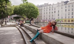 Actor leans back on bank of River Seine wearing short orange dress and knee-high green boots to take a selfie with a bottle of rose wine beside her