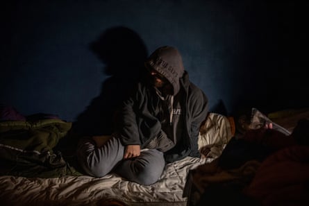 A young man wearing a hoodie sits on a bed in darkness