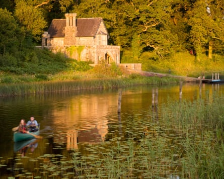 Two people canoe on still waters in evening sunlight past an old stone boathouse