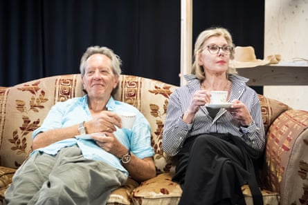 a man and woman sit side by side on a vintage sofa, sipping tea