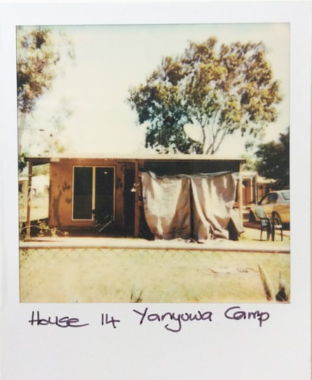 A Polaroid photo of a single-storey house. There are two cloths hanging down from the roof on one side covering the house. There is a mesh fence in the foreground and a tall tree behind the house