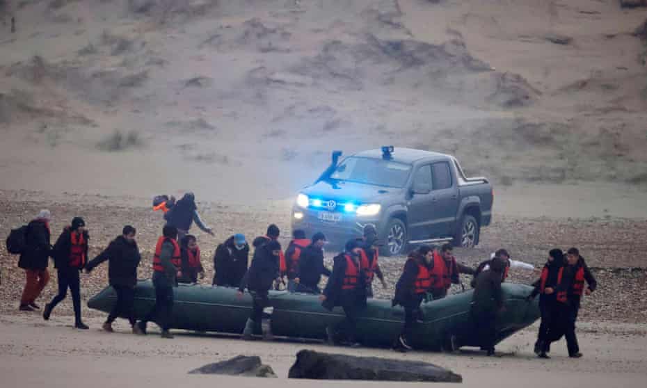 A group of people in red jackets walk a large inflatable boat down a beach. A police vehicle with its blue lights flashing is in the background